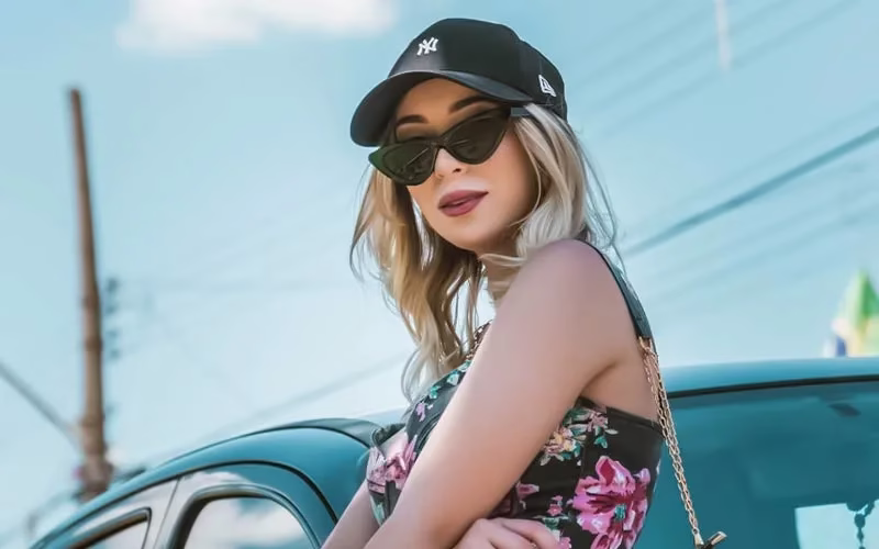 Young woman in floral dress and black cap poses confidently by a car. She wears sunglasses and smiles slightly, exuding a relaxed, stylish vibe, The Culture of the United States: A Complete Guide to the American Lifestyle
